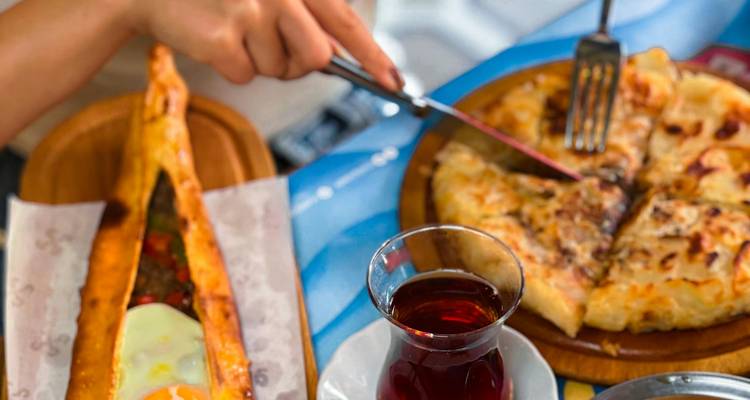 Tableau avec des plats du petit-déjeuner turc : pide à l'œuf, pâtisserie au fromage, et un verre de thé noir tandis qu'une main coupe une tranche.