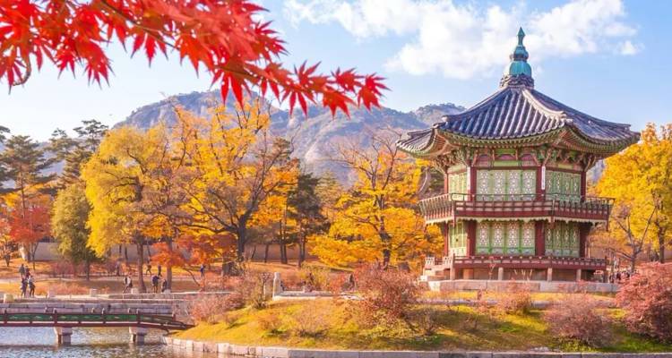 A colorful pavilion sits beside a pond framed by fiery autumn leaves and distant granite peaks.