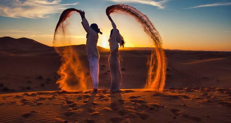 Zwei Personen in traditionellen Gewändern spielen bei Sonnenuntergang in den Sanddünen.
