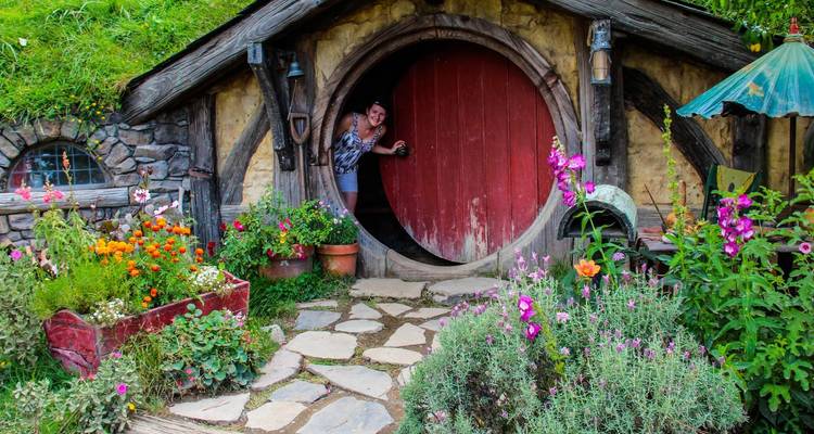 Besucher, der durch eine runde Holztür von Hobbiton blickt, die von farbenfrohen Blumengärten und rustikalen Steinmauern eingerahmt wird.