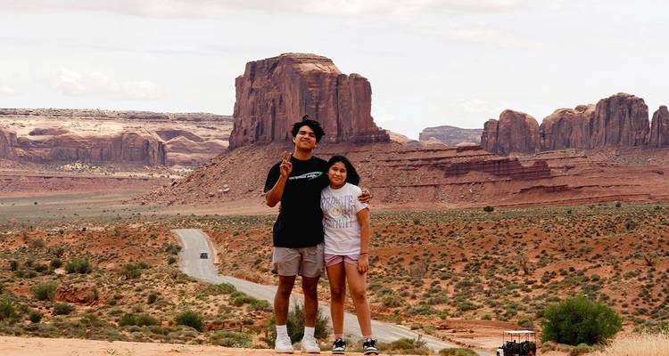 Dos viajeros posan frente a enormes formaciones rocosas rojas del desierto y mesetas bajo cielos brumosos.
