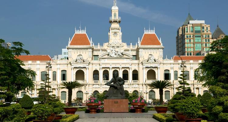 Grand colonial city hall building framed by manicured greenery and a central statue under clear skies