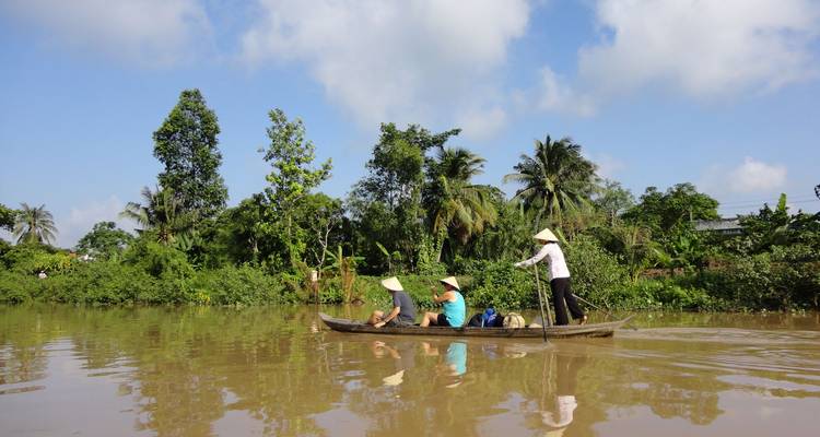 Petit canoë en bois avec des voyageurs glisse le long d'un chenal boueux du Mékong bordé d'une végétation tropicale dense sous un ciel lumineux.