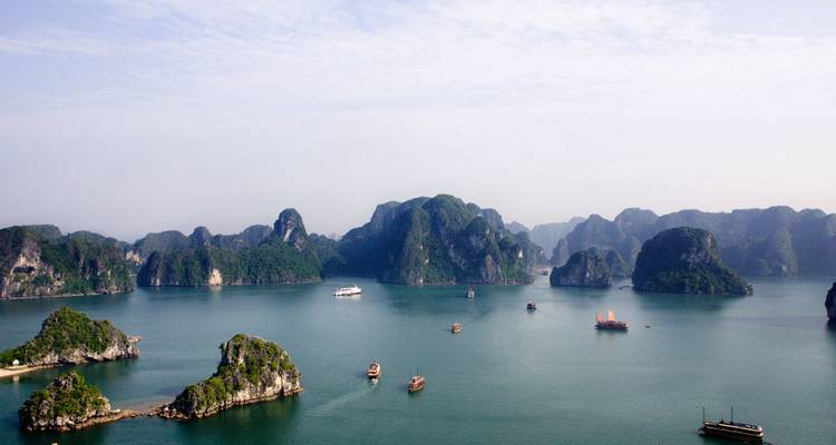 Vue panoramique large des eaux émeraude de la baie d'Halong parsemées d'îlots calcaires et de bateaux de croisière sous un ciel brumeux.