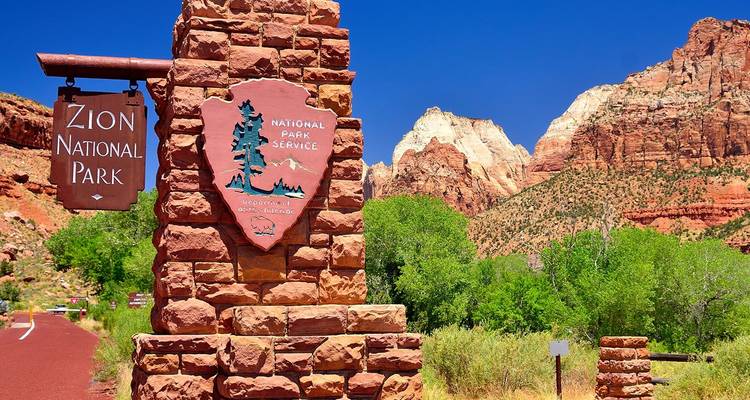 Panneau d'entrée en pierre portant l'inscription Zion National Park devant des falaises rouges et un ciel bleu profond.