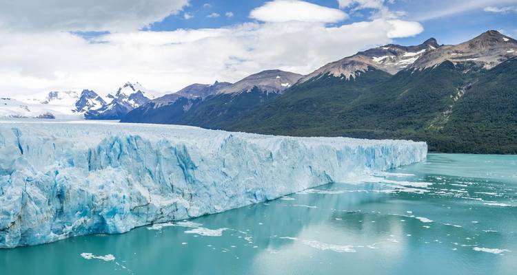 Massive turquoise face of Perito Moreno Glacier meeting calm icy waters with Andean peaks beyond.