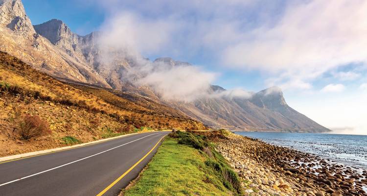 Scenic coastal road with mountains and the ocean beside it under a misty sky.