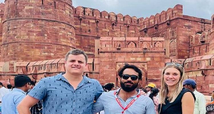 Trois touristes souriants devant les imposants murs de grès rouge du Fort d'Agra au milieu d'une cour animée.