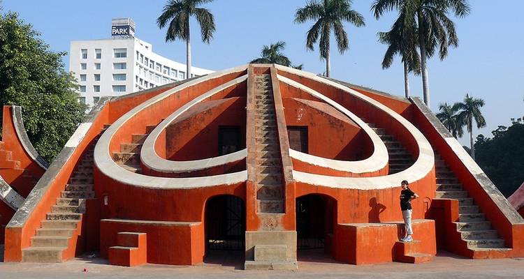 Structure géométrique en pierre rouge de l'observatoire Jantar Mantar à New Delhi encadrée par des palmiers.