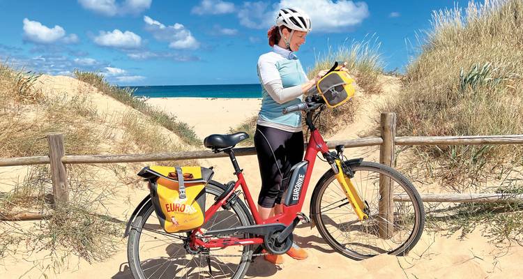 Radfahrer auf einem sandigen Strand mit Dünen und blauem Himmel.