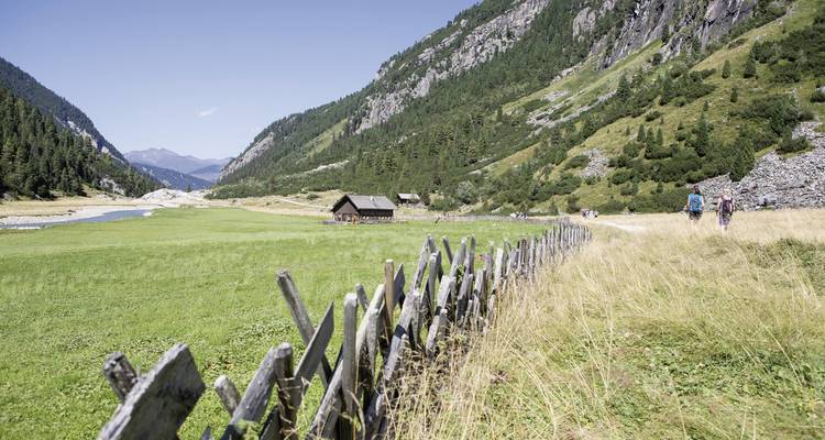 Randonneurs dans une vallée alpine avec des clôtures en bois et des prairies.