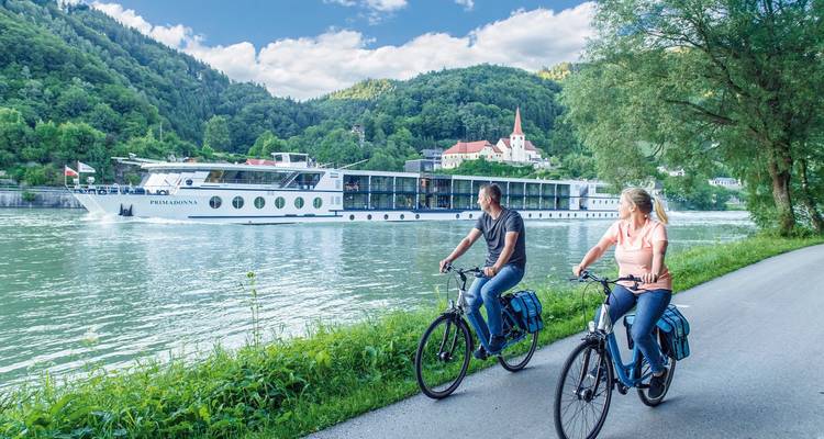 Radfahrer bei einer Kirche und einem Fluss mit einem Kreuzfahrtschiff im Hintergrund.