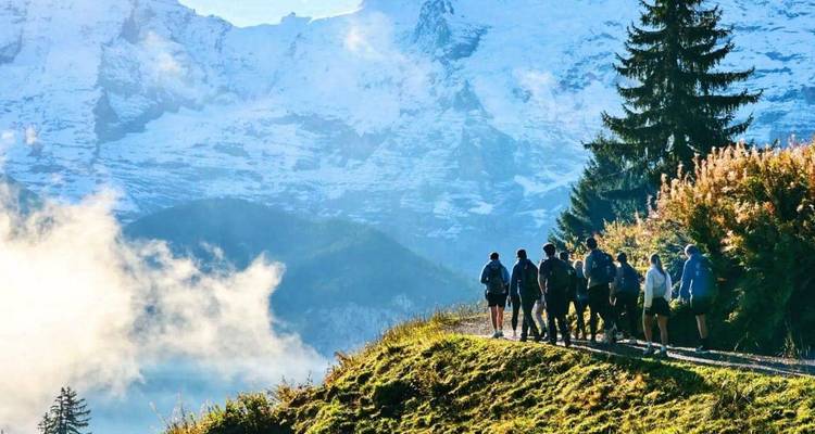 Group of trekkers on a mountain trail with dramatic snowy Swiss peaks and swirling clouds behind.