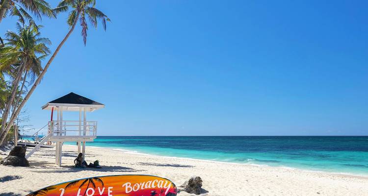Unberührter Strand mit weißem Sand und türkisfarbenem Wasser, ein Rettungsschwimmerturm und schräg stehende Palmen unter einem klaren blauen Himmel; ein farbiges Surfbrett mit der Aufschrift "I LOVE Boracay" im Vordergrund.