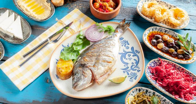 Grilled whole fish meal accompanied by colourful mezze dishes on rustic painted wooden table.