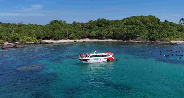Een speedboot voor anker in turquoise water bij een klein tropisch eiland waar snorkelers zwemmen.