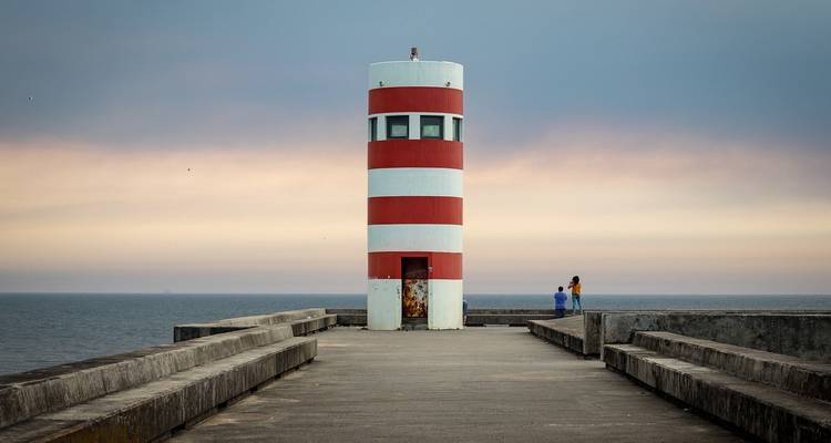 Gestreifter Leuchtturm auf einem Pier mit Meerblick und zwei gehenden Personen.