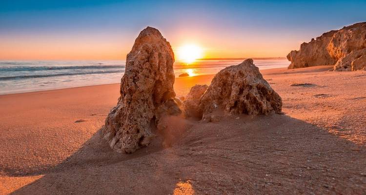 Sonnenuntergang an einem Strand mit Felsformationen und einem ruhigen Meer.