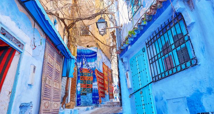 Rue étroite avec des bâtiments peints en bleu et des tapis suspendus.