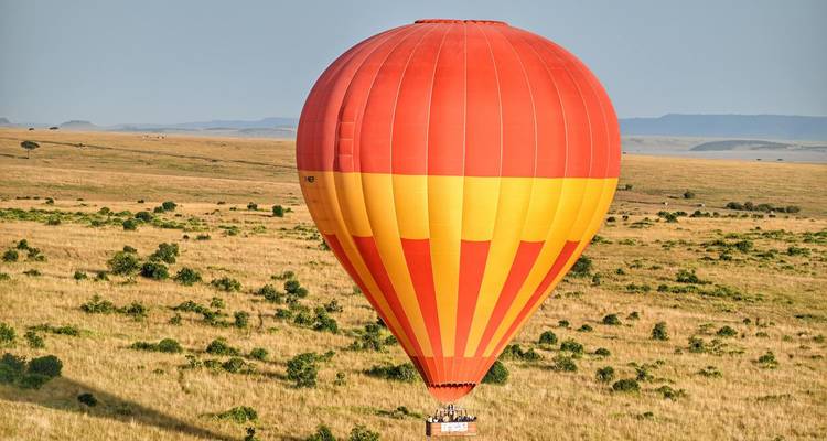 A vibrant red-and-yellow hot-air balloon floats over golden grass plains with passengers peering out.