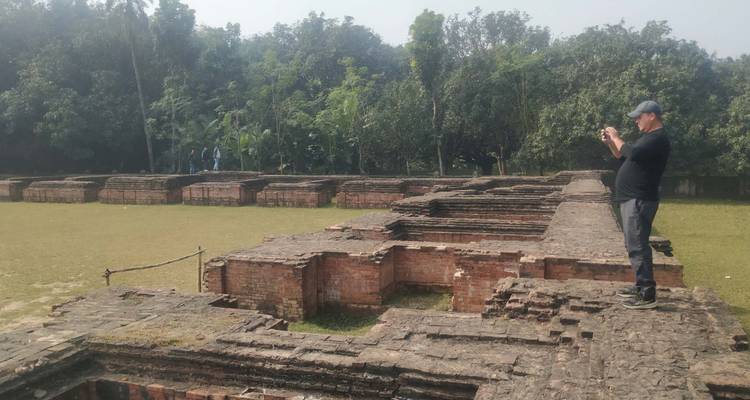 A tourist photographs low brick ruins against a backdrop of dense greenery.