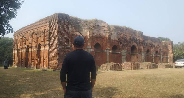 Back view of a man observing a historic red-brick mosque surrounded by dry grass.