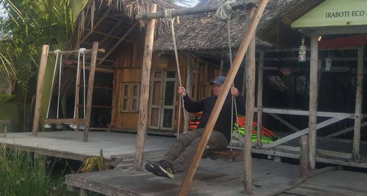 Un hombre se relaja en un columpio de madera frente a cabañas ecológicas con techo de paja junto al agua.