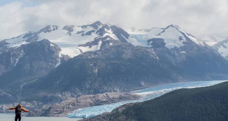 Einzelne Wanderin erhebt die Arme auf einer Klippenkante mit Blick auf ein von Gletschern ausgehöhltes Tal mit aufragenden verschneiten Bergen