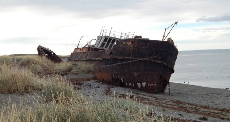 Verrostetes Schiffswrack ruht auf einem windigen Sandstrand mit hohen Gräsern und grauem Meer dahinter