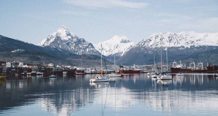 Der Hafen von Ushuaia mit vertäuten Segelbooten auf spiegelglatter See, die schneebedeckte Gipfel unter einem blassen blauen Himmel widerspiegelt