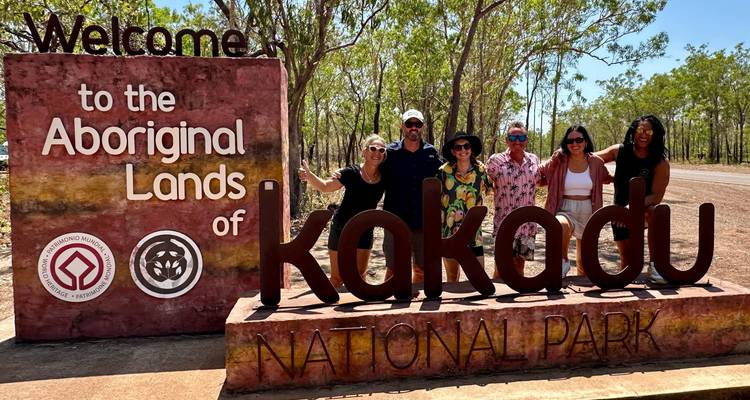 Des voyageurs souriants posent devant le panneau de bienvenue du parc national de Kakadu sous les eucalyptus et un ciel bleu.