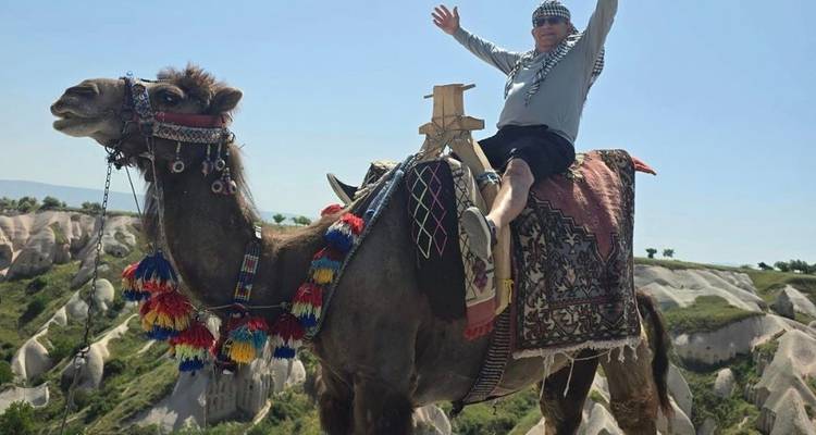 Persona montando un camello con formaciones rocosas panorámicas detrás.