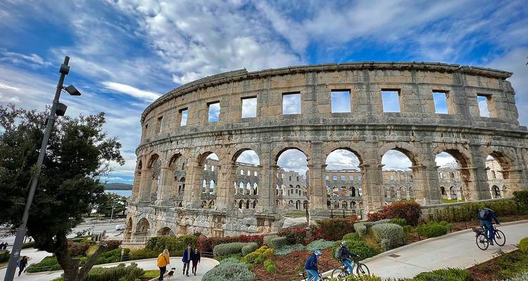 Amphithéâtre romain bien préservé à Pula sous un ciel dramatique avec des visiteurs et des cyclistes à proximité.