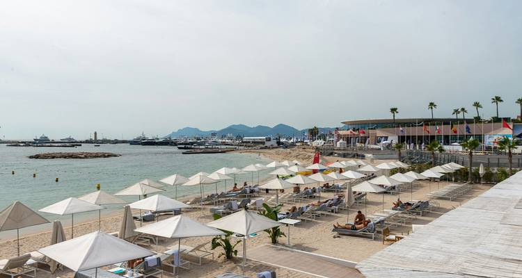 Breiter Sandstrand in Cannes mit Sonnenliegen und Sonnenschirmen, der eine ruhige Bucht und entfernte Berge überblickt.
