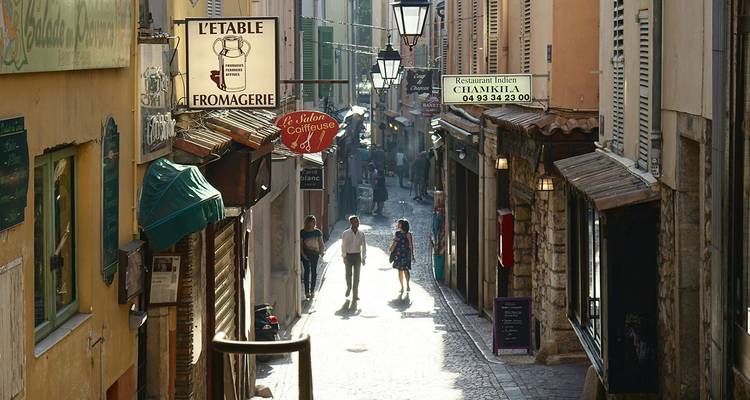 Enge Fußgängerstraße in Antibes mit Cafés, Schildern für eine Käserei und Restaurants sowie einem Paar, das am Nachmittag spaziert.