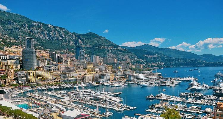 Luftaufnahme der dichten Skyline von Monaco, des segelbootgefüllten Yachthafens und der steilen Berge, die sich hinter der Stadt erheben.