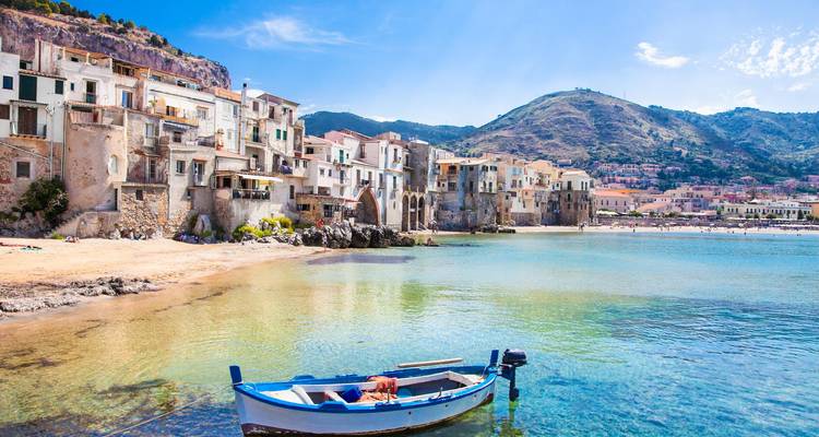 Picturesque seaside village of Cefalù with pastel houses lining a tranquil bay and a blue boat in the foreground.