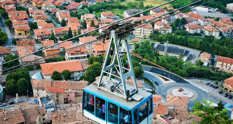 Blue cable car climbing above tiled rooftops toward the mountaintop of San Marino.