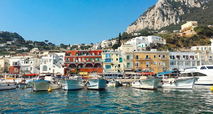 Colorful waterfront buildings and moored boats in Capri’s bustling harbor beneath a towering limestone cliff.