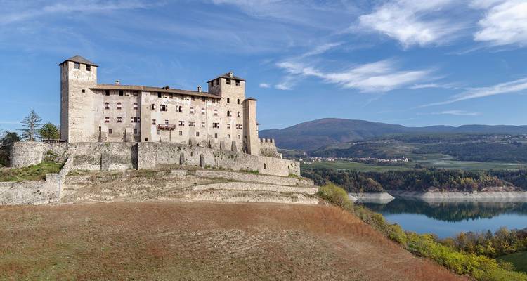 Un château de pierre médiéval couronne une colline surplombant un lac bleu avec des montagnes boisées au-delà.