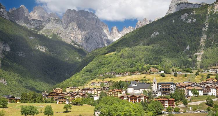Un village alpin se situe dans une vallée verdoyante et luxuriante sous les pics dentelés des Dolomites et un ciel bleu éclatant.