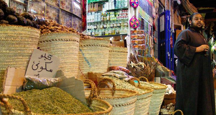 Canastas coloridas llenas de especias y hierbas secas llenan un puesto animado del mercado interior mientras un hombre con ropa tradicional se para a su lado