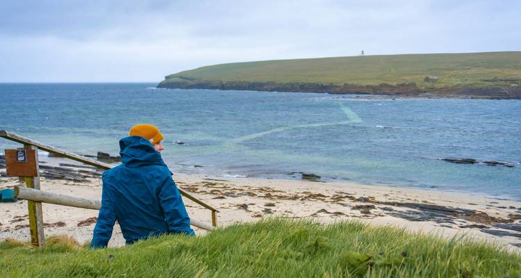 Ein einzelner Reisender in einer gelben Mütze blickt von grasbewachsenen Dünen auf einen windigen Strand und eine aufgewühlte See.