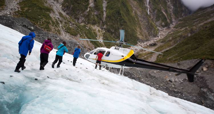 Aventureros con chaquetas de colores caminan a través de una superficie glacial hacia un helicóptero que los espera cerca.