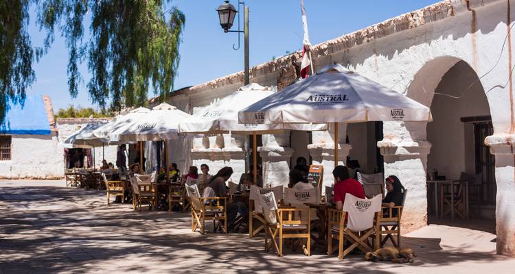Les voyageurs se détendent sous des parasols blancs dans un café rustique en plein air situé sur une place bordée de maisons en adobe dans un village désertique.