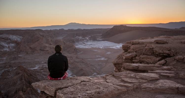 Un voyageur solitaire assis sur une corniche rocheuse dominant le désert otherworldly de la Valle de la Luna au coucher du soleil.