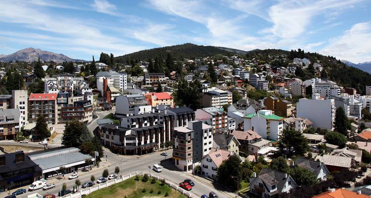 Vista panorámica elevada de los barrios residenciales de Bariloche enmarcados por colinas cubiertas de pinos.