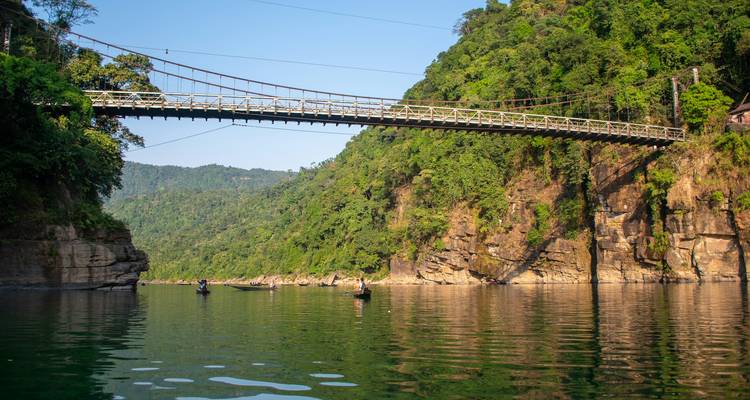 Een hangbrug over een rivier met groene heuvels op de achtergrond.