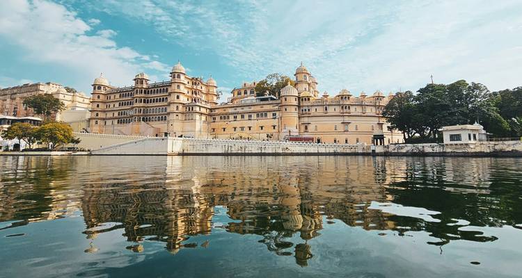 Udaipur City Palace reflected in still waters under a bright sky.