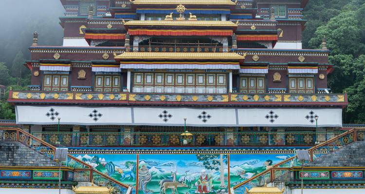 Une façade de monastère bouddhiste himalayen richement décorée brille avec des rouges, des ors et des bleus vibrants sur fond de collines verdoyantes luxuriantes.